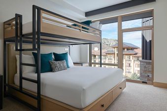 A modern bunk bed with a queen-sized bed on the bottom and a twin on top, featuring white linens and teal pillows, beside a large window overlooking mountains and resort buildings.