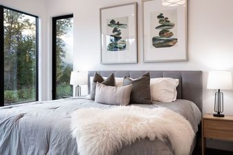 A bedroom with a large bed, two windows that show a view of trees outside, and two pieces of artwork depicting abstract shapes in the form of stacked stones.