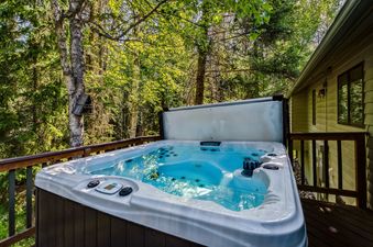 A private outdoor hot tub with bright blue water is set on a wooden deck, surrounded by lush green trees and a rustic bird feeder, with a glimpse of the house in the background.