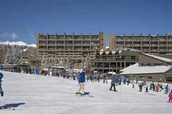 A wide view of the One Bedroom Suite Beaver Run Resort from the ski slopes, showing skiers of all ages, the large resort buildings, and snow-covered mountains under a bright blue sky.