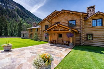 The Elk Mountain Lodge is a rustic log cabin nestled against a lush mountain landscape with a clear blue sky overhead.