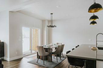 A modern dining area in The Circle Unit B features a dark wood table with eight upholstered chairs, illuminated by an elegant black chandelier. Adjacent is a contemporary kitchen island with seating and sleek pendant lights, all set against light-colored