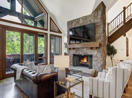 A cozy living room at Snow Cloud Lodge 715 with a stone fireplace, television, brown leather sofa, striped armchairs, and large windows looking out to a lush forest.