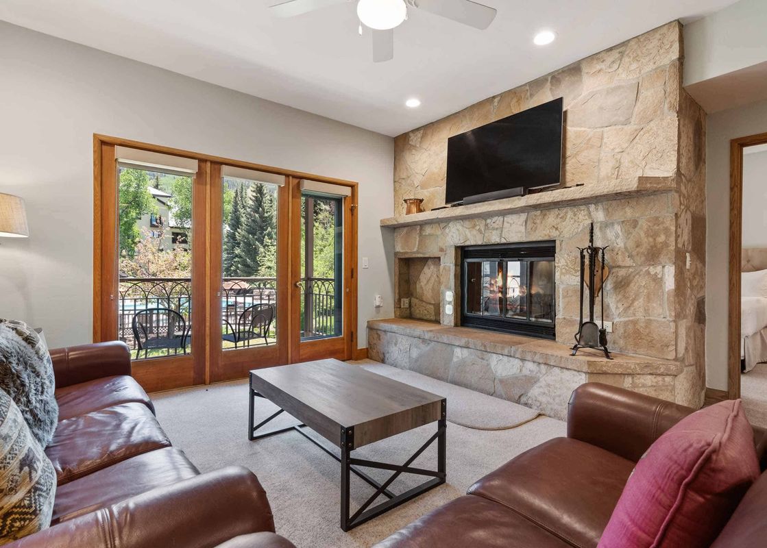 A cozy lounge area with brown leather sofas, a modern coffee table, and a large stone fireplace with a mounted television, opening to a balcony with green trees visible.