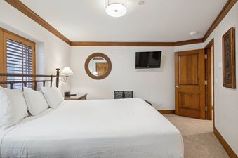 The primary bedroom at The Charter L032 features a bed with white linens, a bedside table with a lamp, a wall-mounted TV, and a round mirror with a woven frame.