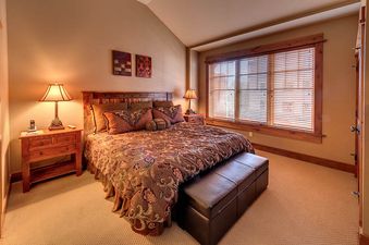 A bedroom with a king size bed, a large window with blinds, and a leather ottoman.