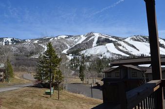 A wide view from the Ranch at Steamboat RA405, showing snow-capped mountains with ski slopes, green pine trees, and a winding road through residential buildings under a blue sky.