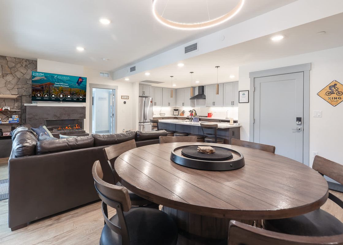 A modern and inviting open-concept living space in The Aerie, featuring a large television above a fireplace, a comfortable leather sectional, and a circular dining table, all seamlessly connected to a contemporary kitchen.