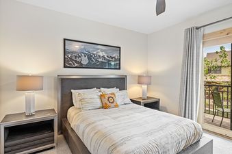 A cozy bedroom with a king-sized bed, two nightstands with lamps, a mountain art piece above the bed, and a balcony with an outdoor view.
