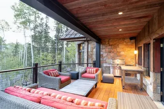A view of a wooden deck with wicker patio furniture, a gas grill, and a wooden table, all overlooking a green forest with a wood-paneled ceiling above.