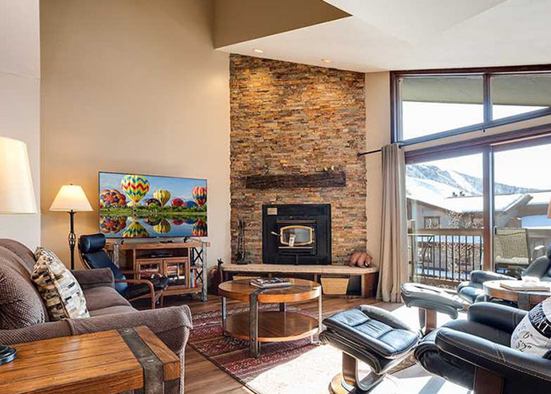 The living room of the Ranch at Steamboat RA106 features a brown sofa, two black recliners, a coffee table, a TV with hot air balloons, a stone fireplace, and large windows with a mountain view.