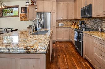A modern kitchen with granite countertops, light wood cabinetry, hardwood floors, stainless steel appliances, and a window with a view of green trees.