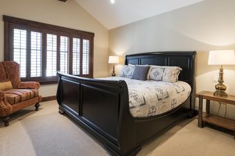The primary bedroom features a black sleigh bed with patterned bedding, two nightstands with lamps, a rust-colored armchair, and large windows with dark wood shutters.