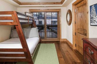 A bedroom in Vail Mountain View 402 features a bunk bed with a full-size bed on the bottom and a twin-size bed on the top. The room has a wooden floor, a green rug, and a window with a view of the mountains.
