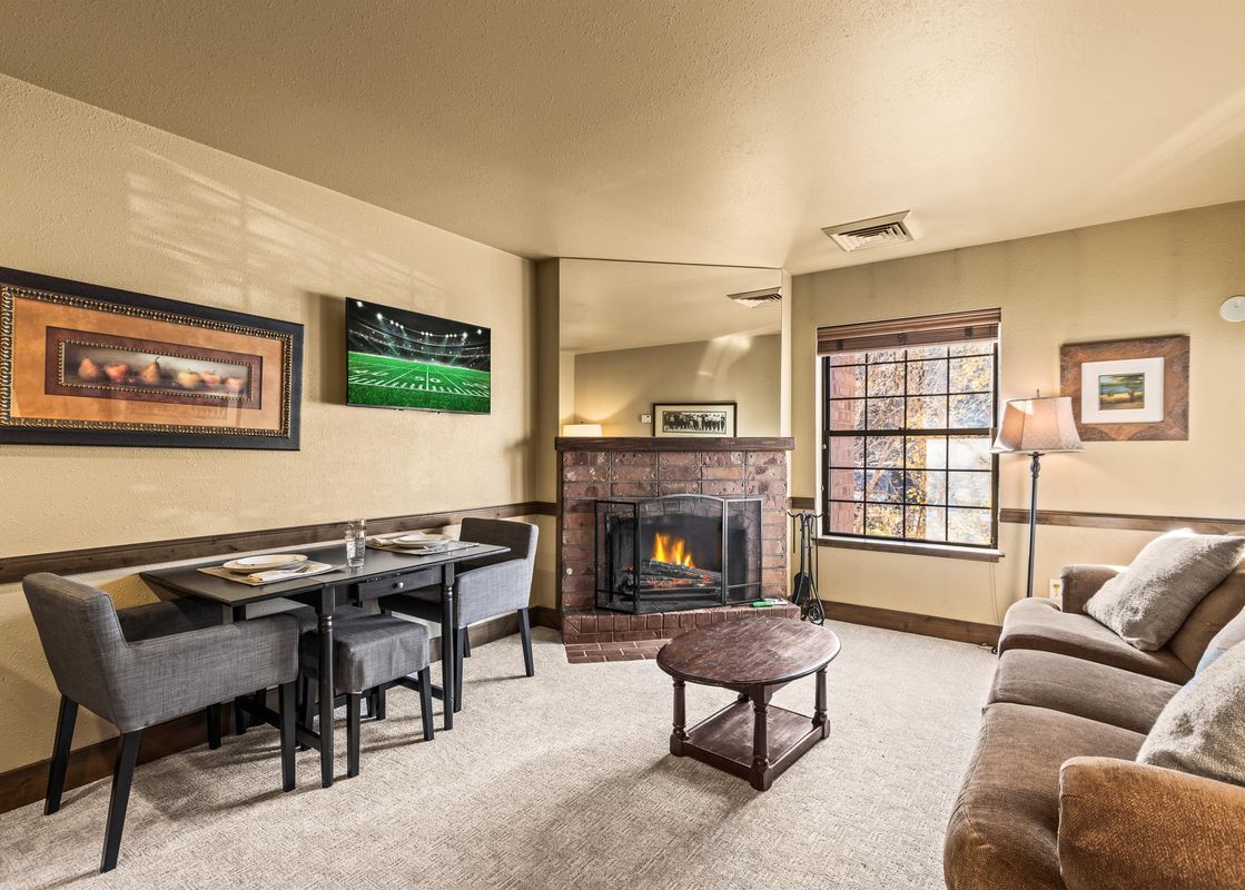 A cozy living and dining area at Copperbottom Inn 103 featuring a brick fireplace, a dining table with four chairs, a sofa, and a large window.