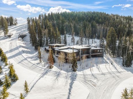 An aerial view of the Quicksilver Haus, a modern multi-level building, surrounded by snow-covered pine trees and a vast snowy landscape under a clear blue sky.