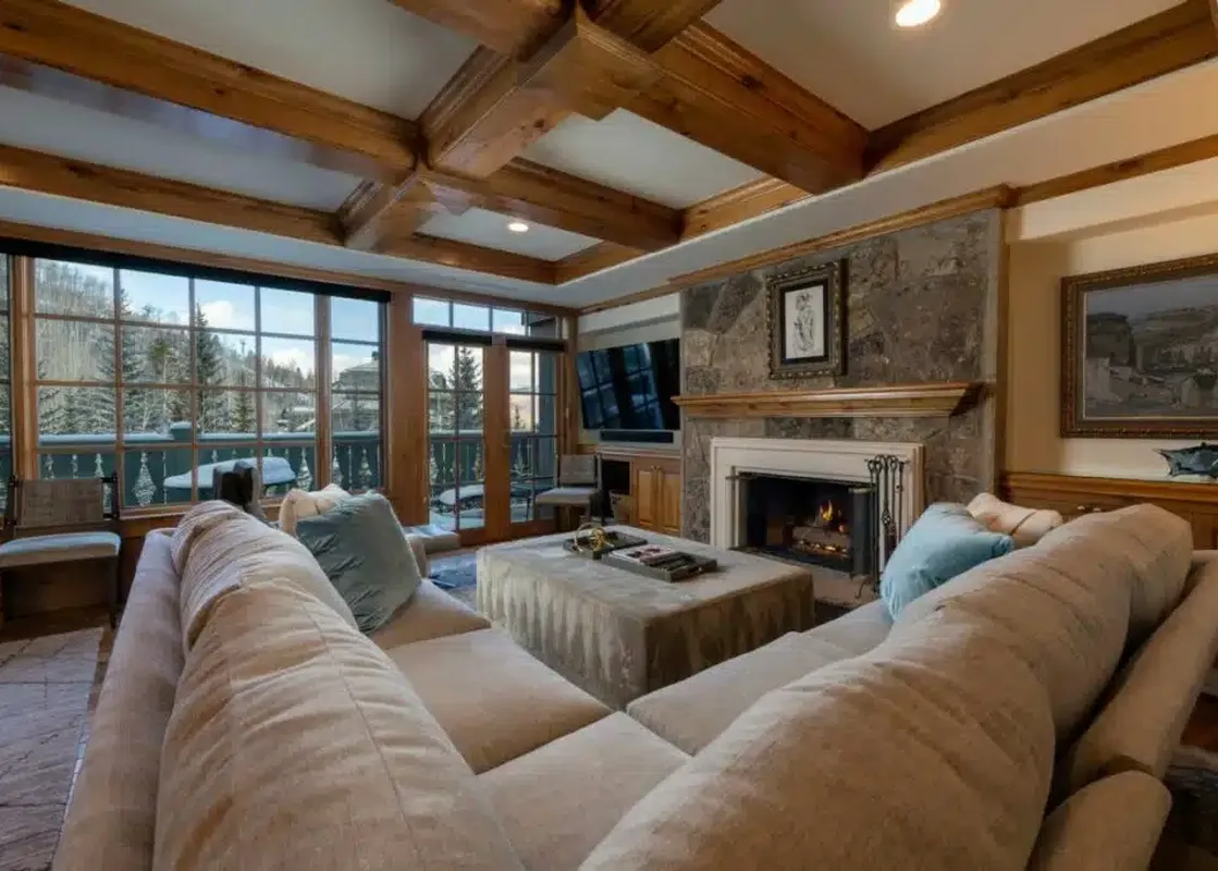 A cozy living room with a large sectional sofa, a fireplace set in a stone wall, and expansive windows offering a winter mountain view.