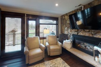 A cozy living room with a fireplace and a TV, two armchairs, and a balcony with a view of the mountains