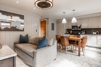 A comfortable open-concept living room and kitchen at Cedar Crest Chickadee Cottage 2, featuring a gray sofa, a wooden dining table with leather chairs, and modern kitchen appliances.