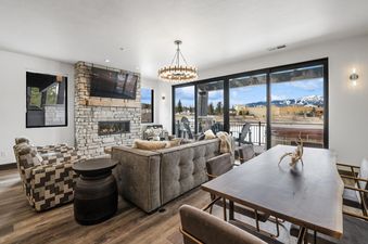 A spacious open-concept living area at Huckleberry Haven with a fireplace, comfortable seating, and a dining table, featuring large windows that offer stunning mountain views.