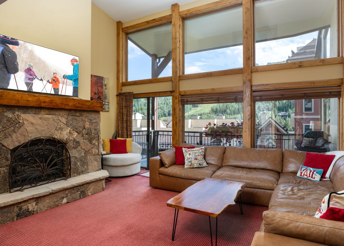 A spacious living room at Montaneros 417 featuring a large leather sofa, a unique wooden coffee table, a stone fireplace with a television above it, and wide windows with mountain views.