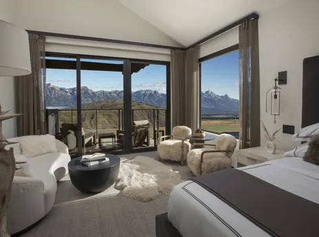 A luxurious bedroom in the Grand Teton Suite features a large bed, plush seating, and expansive windows offering stunning views of the mountains.