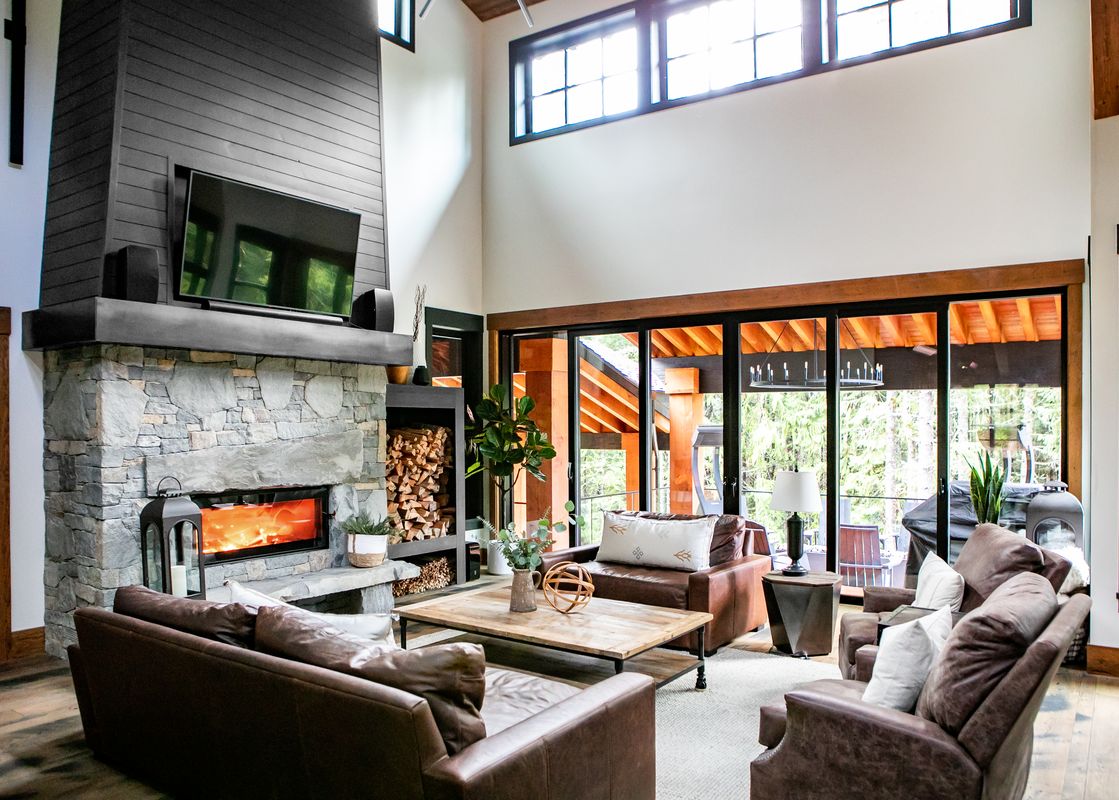 A cozy living room in Falcon Cottage featuring a stone fireplace with a roaring fire, a TV, and comfortable leather seating arranged around a wooden coffee table, with a large glass door opening to a balcony and a forest view.