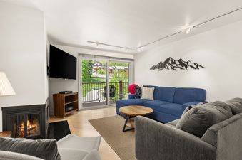 A modern and cozy living room featuring a blue sectional sofa, a grey armchair, a wooden coffee table, a flat-screen TV, a fireplace, and a balcony with an exterior view.