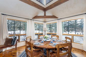 A wooden dining table with six chairs is set in a dining room with floor-to-ceiling windows offering views of a snowy landscape.