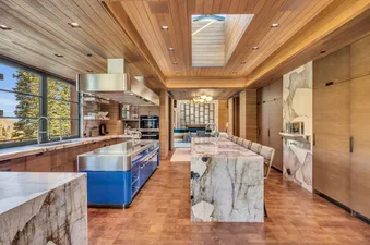 A modern kitchen at Monitors Rest, featuring wooden ceilings, large windows with outdoor views, and two central islands, one blue and stainless steel, the other a long marble slab.