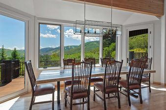 A spacious dining room at Timer Ridge Retreat features a long wooden table with rustic chairs, bathed in natural light from large windows offering panoramic mountain views and direct access to a balcony.