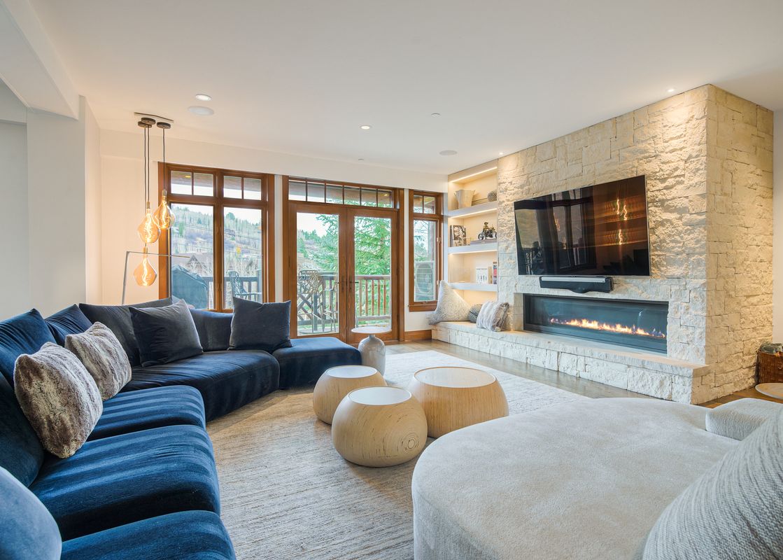 A large blue sectional sofa and unique round coffee tables in the lounge at Snow Cloud Lodge 509, with a fireplace and television on a stone wall, large windows overlook a green outdoor scene.