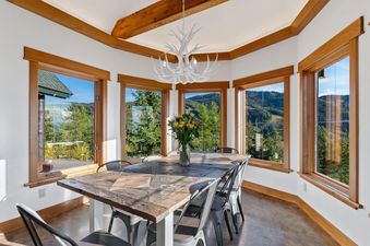 The dining area at Ridge Top Chalet features a rustic wooden table and industrial-style chairs, surrounded by large windows showcasing panoramic mountain and forest views.