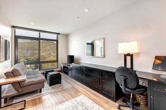 A modern living area in Lowell 402B features a grey futon, a long black storage console with a TV and a workspace, and large windows offering a scenic mountain view.