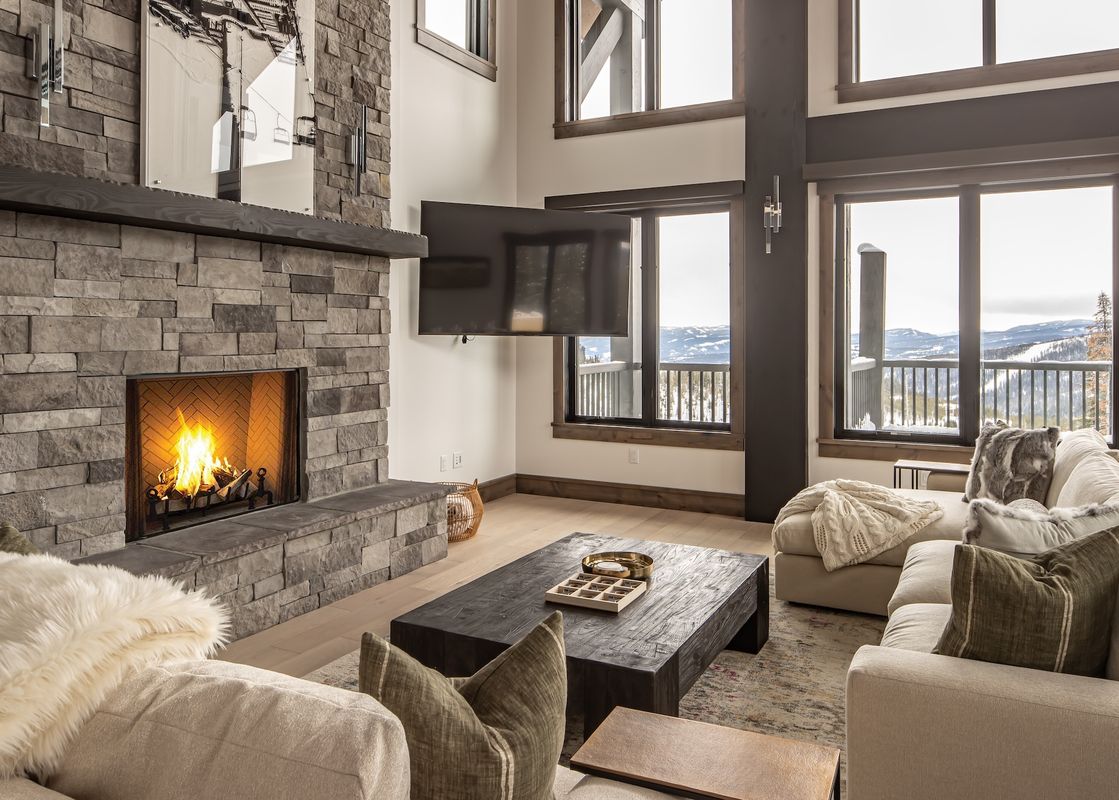 The living area at 22 Lower Cascade Ridge features a stone fireplace, a large flat-screen television, and expansive windows that offer stunning views of the snow-covered mountains.