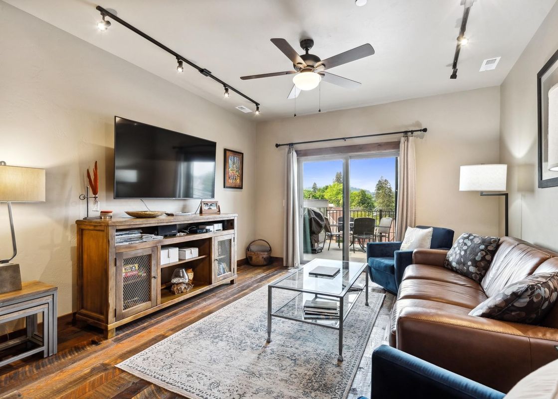 A cozy living room in Baker Loft 302 featuring a large TV, a brown leather sofa, blue armchairs, and a glass coffee table, with a balcony offering a view of lush greenery.