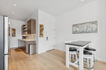 A modern kitchen with stainless steel appliances and brown cabinets, adjacent to a dining area with a high-top table and three bar stools.