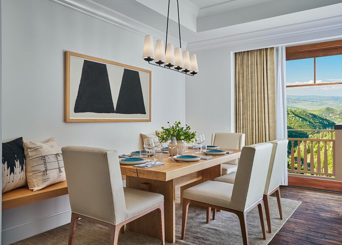 The dining area in the Peak View Two Bedroom Residence features a wooden dining table set with place settings and a decorative vase, surrounded by light-colored chairs and a built-in bench.