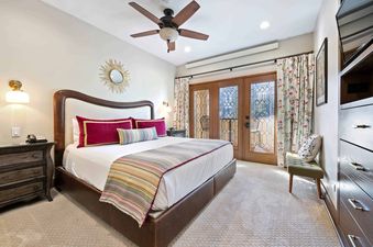 A king-sized bed with a leather headboard and colorful bedding is in the center of the bedroom, with French doors leading to a balcony on the right and a dresser with a television on the left.