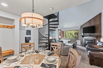 A spacious dining area with a round wooden table set for a meal, a black spiral staircase leading upstairs, and a living room with comfortable seating, a fireplace, and large windows looking out to green trees.