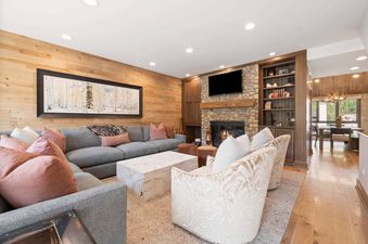 The inviting living area features a stone fireplace, a large gray sectional sofa with pink accent pillows, and light wood paneled walls.