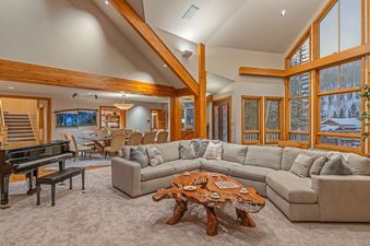 A spacious living room with a large sectional sofa, wooden coffee table, piano, large windows with mountain views, and a dining table in the background.