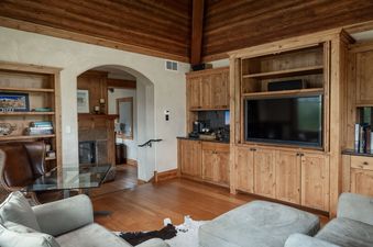 Rustic living room with high wood-paneled ceilings, a built-in media center, cozy seating, and an arched entryway leading to a fireplace.
