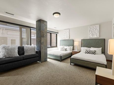 A modern guest bedroom at Lowell 401A featuring two queen beds with green headboards, white bedding, a dark gray sofa with fluffy white pillows, and large windows with blinds.