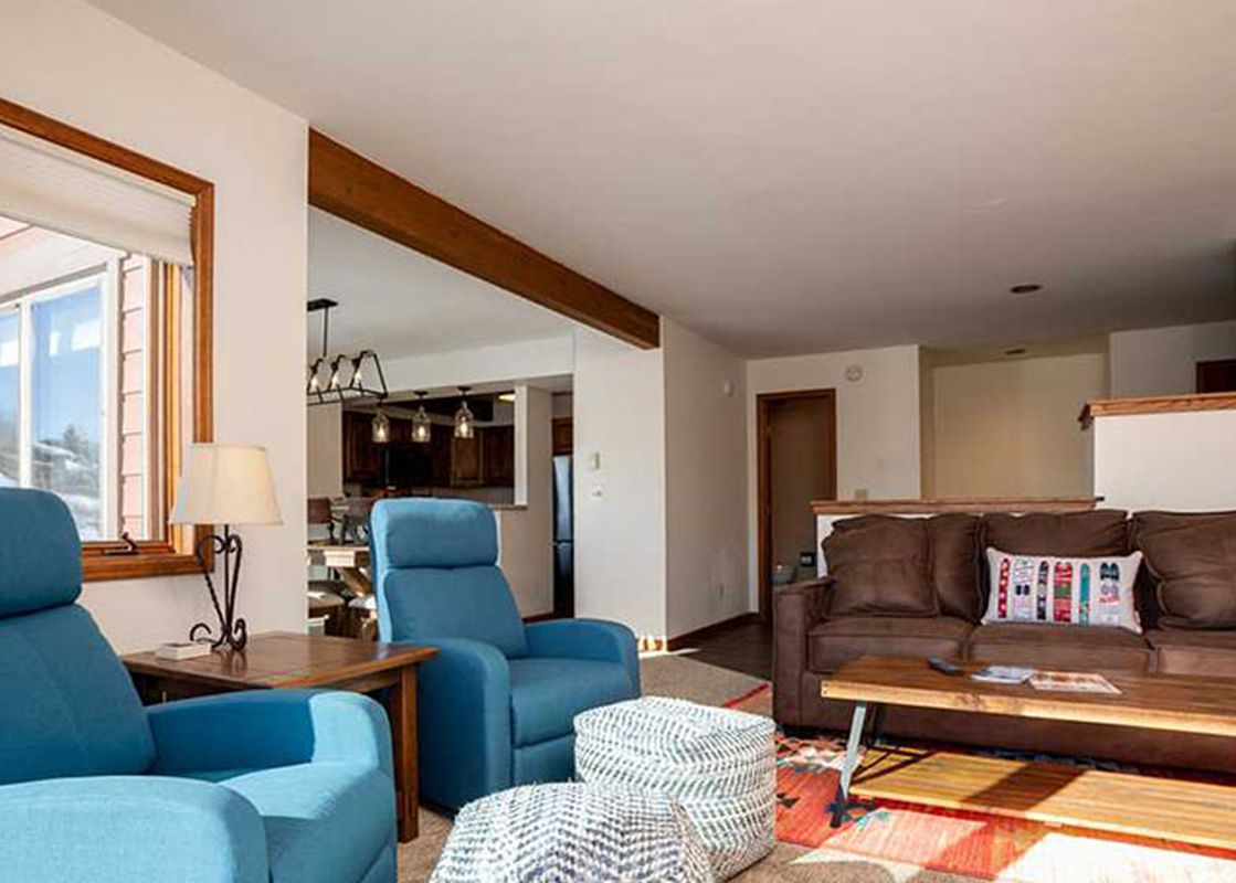 A cozy living room in The Ranch at Steamboat RA507 featuring a brown sofa, blue armchairs, a wooden coffee table, and large windows offering natural light and a view of the surrounding property.