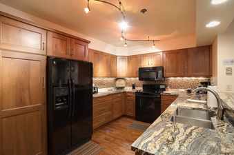 A modern kitchen with dark wood cabinets, a black refrigerator, mosaic tile backsplash, granite countertops, and hardwood floors is illuminated by track lighting.