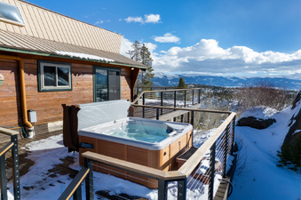 An inviting hot tub on a snow-dusted wooden deck, offering panoramic mountain views under a bright blue sky.