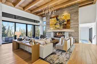 A spacious and modern living room features a stone fireplace, abstract artwork, plush seating, a wooden console table, and large windows with outdoor views.
