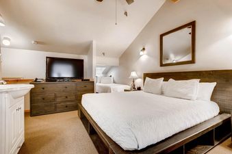 A spacious bedroom at Powder Pointe 303B features two beds with white bedding, a dresser with a flat-screen TV, and a vanity sink, all under angled ceilings.