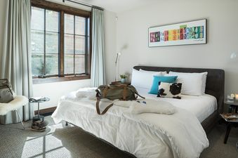 A king-sized bed with white linens, a turquoise pillow, and a cowhide print cushion, accompanied by a large window and a colorful art piece above the bed in Fish Creek Lodge 11.